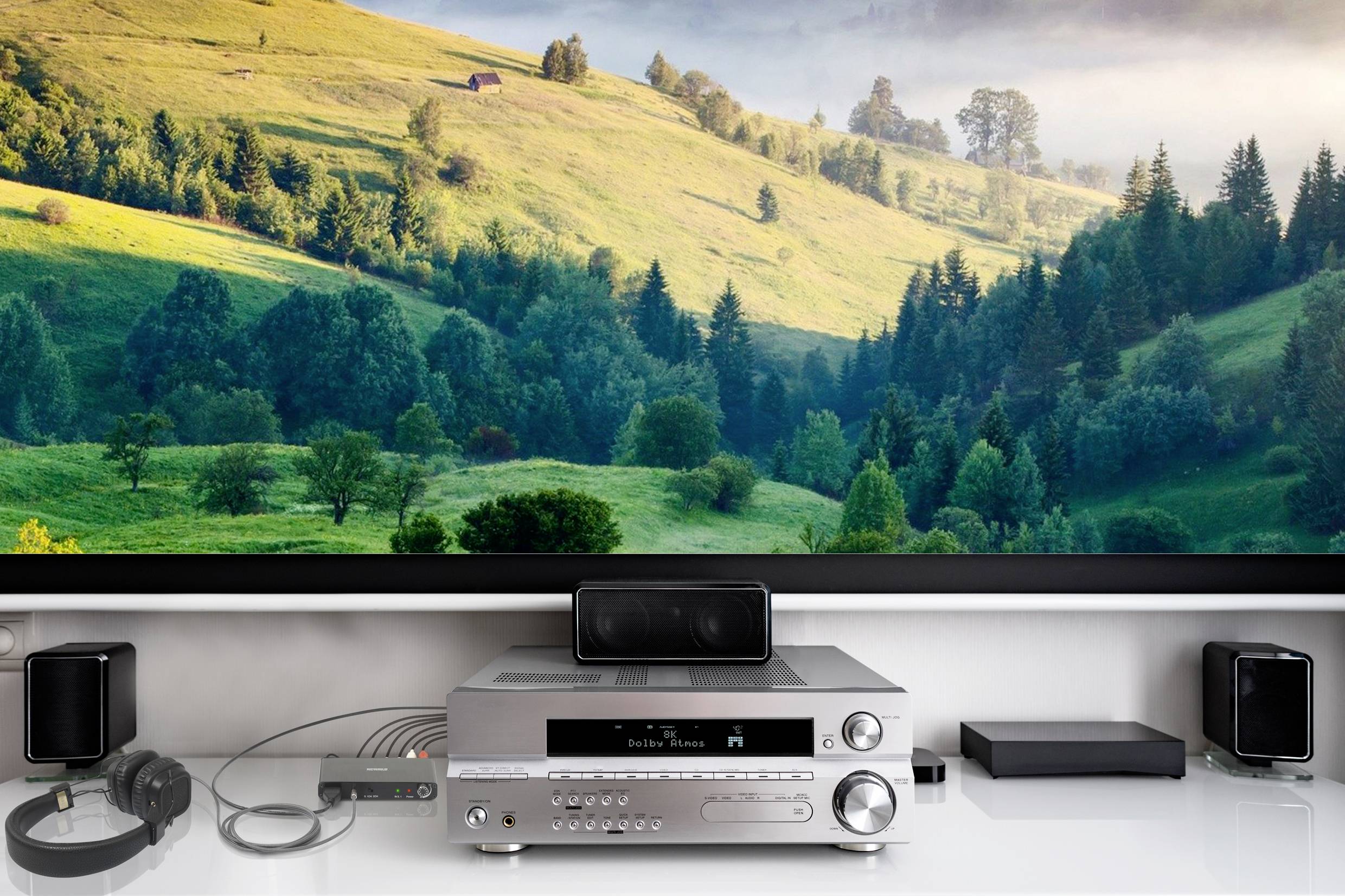 A home cinema system with speakers and an amplifier stands in front of a large screen displaying a woodland and meadow landscape.
