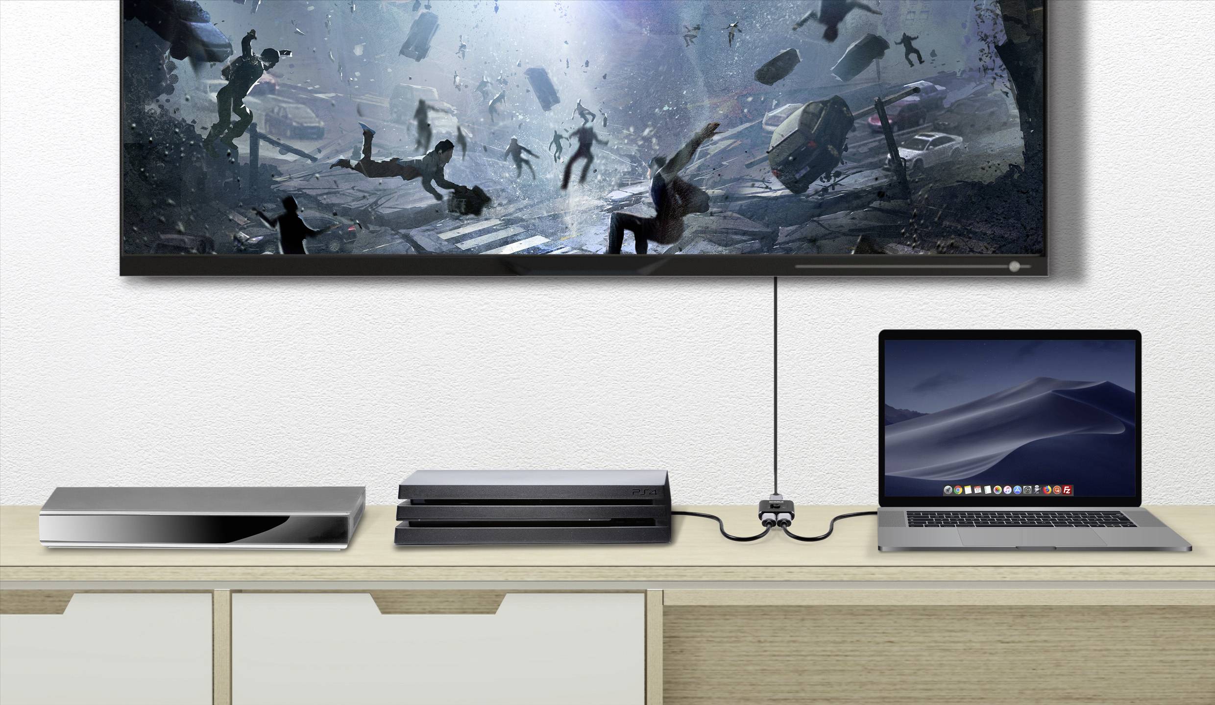 Laptop and consoles on a table in front of a monitor displaying a dynamic film scene with people and debris.