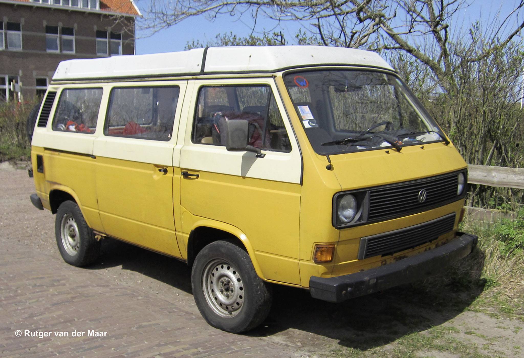 A yellow vintage van is parked on a paved path. Trees and a tiled roof are visible in the background.