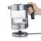 A transparent electric kettle with stainless steel details shows maximum and minimum water level indicators. A hand is grasping the handle.