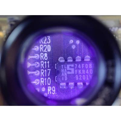 Close-up of a circuit board with a microchip and solder points; the chip is illuminated under purple light and displays PIN labels.