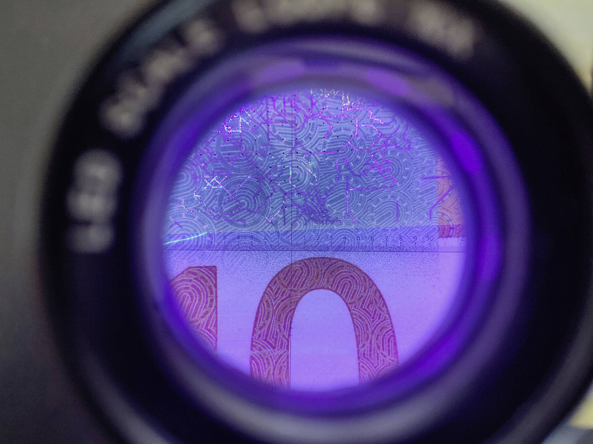 A purple light illuminates security features of a banknote through a magnifying glass to detect counterfeits.