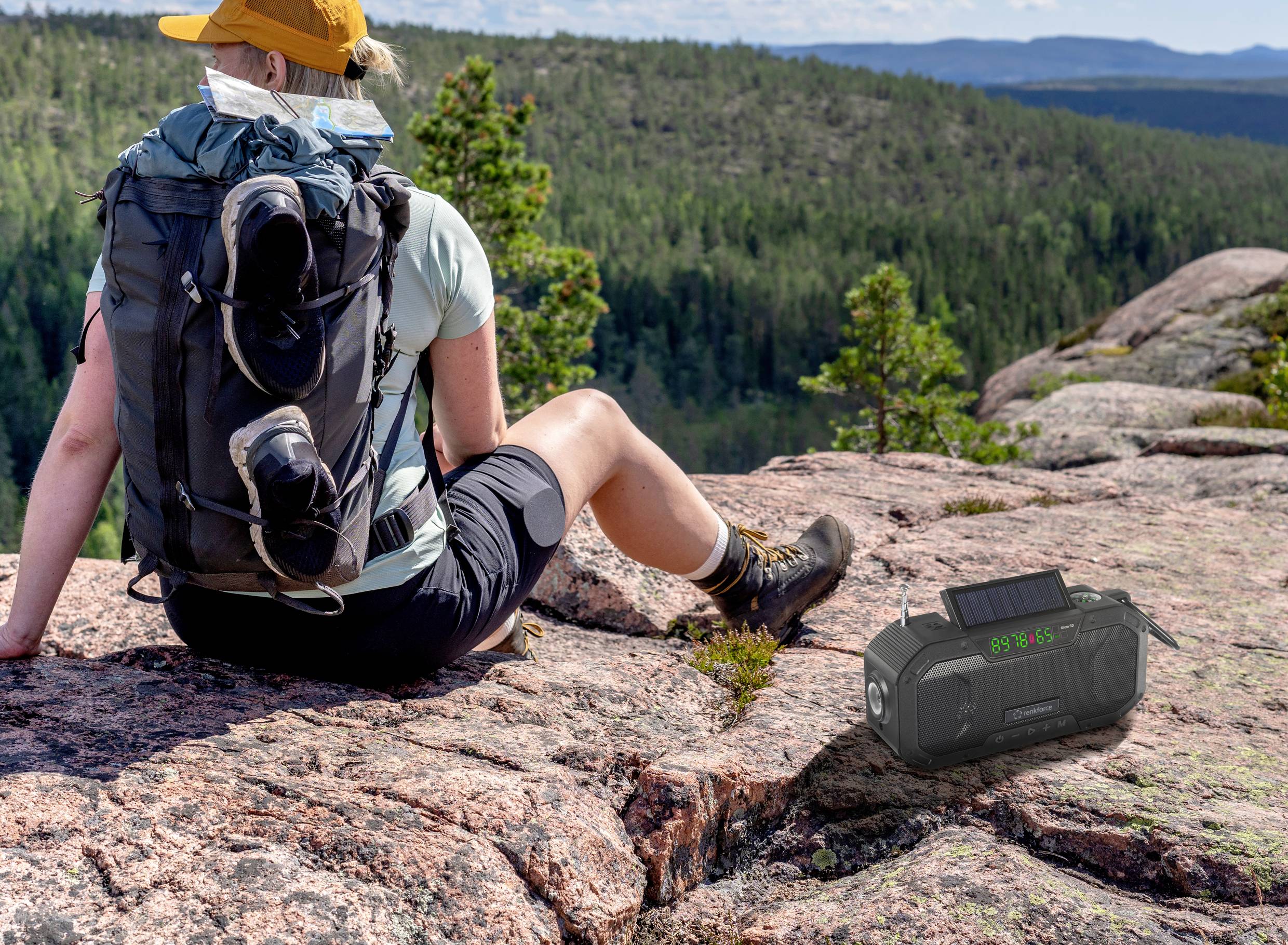A person is sitting on a rock in a mountainous landscape, with a rucksack and a portable music speaker beside them.