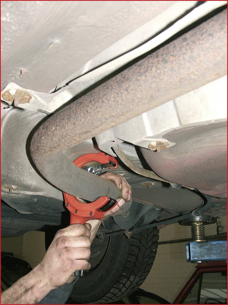 A person is repairing the exhaust of a car in a garage, holding a tool to secure the exhaust underneath the vehicle.