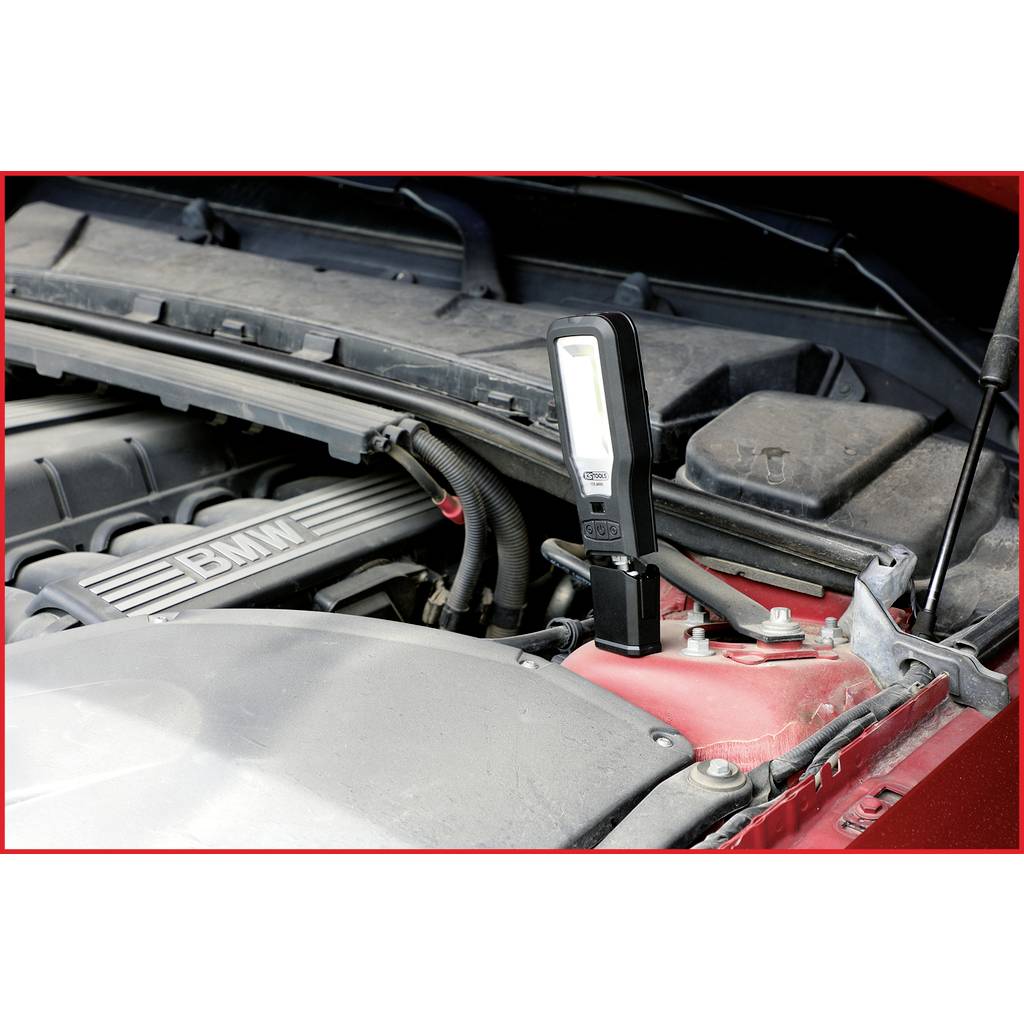 A car engine compartment with a portable lamp directed onto the engine. The cover displays the 'BMW' logo.