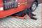 A mechanic is working on a creeper underneath a red van on a paved car park.