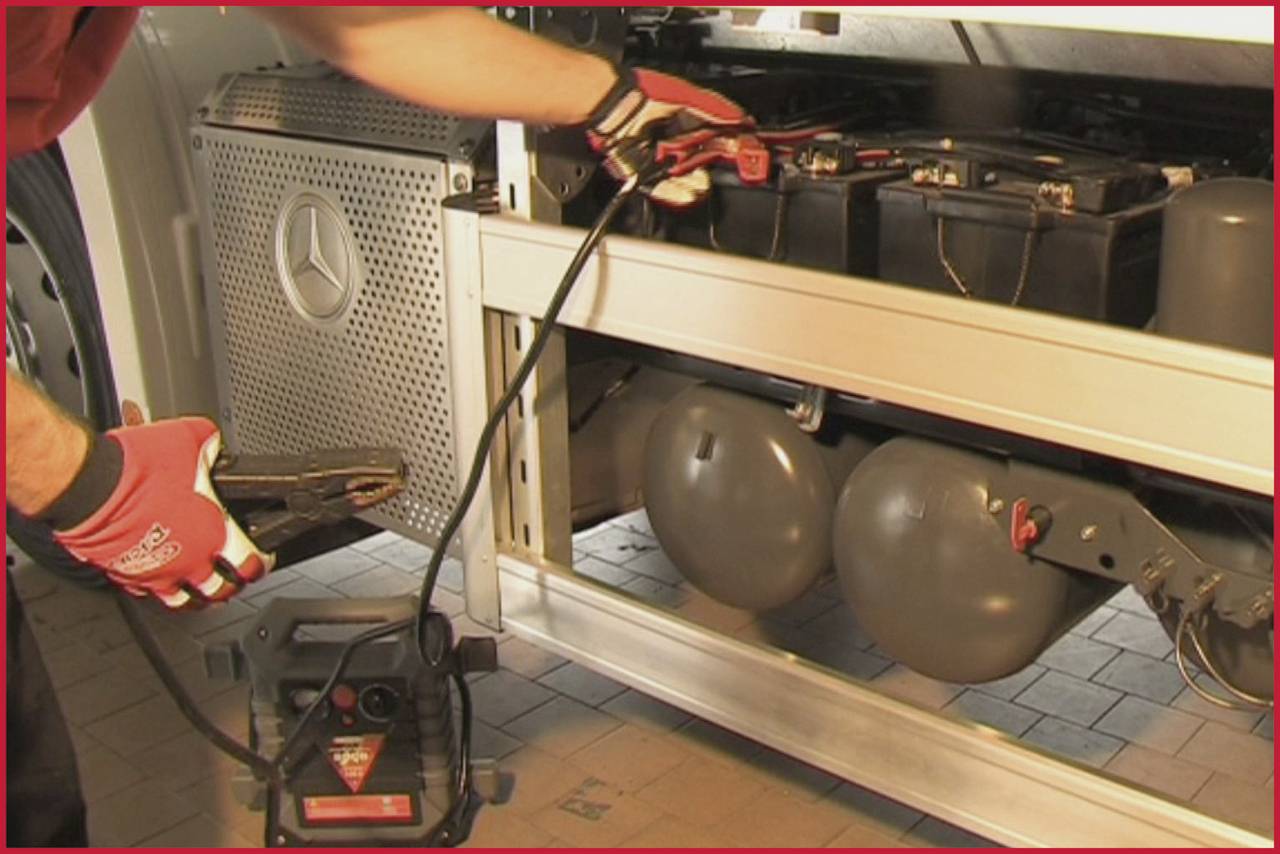 A person connects a jump lead to a battery in a vehicle. Car parts are visible in the background.