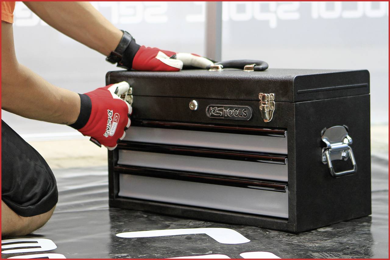 A person is wearing red gloves and opening a black toolbox. The box has multiple drawers and is standing on a floor covering.