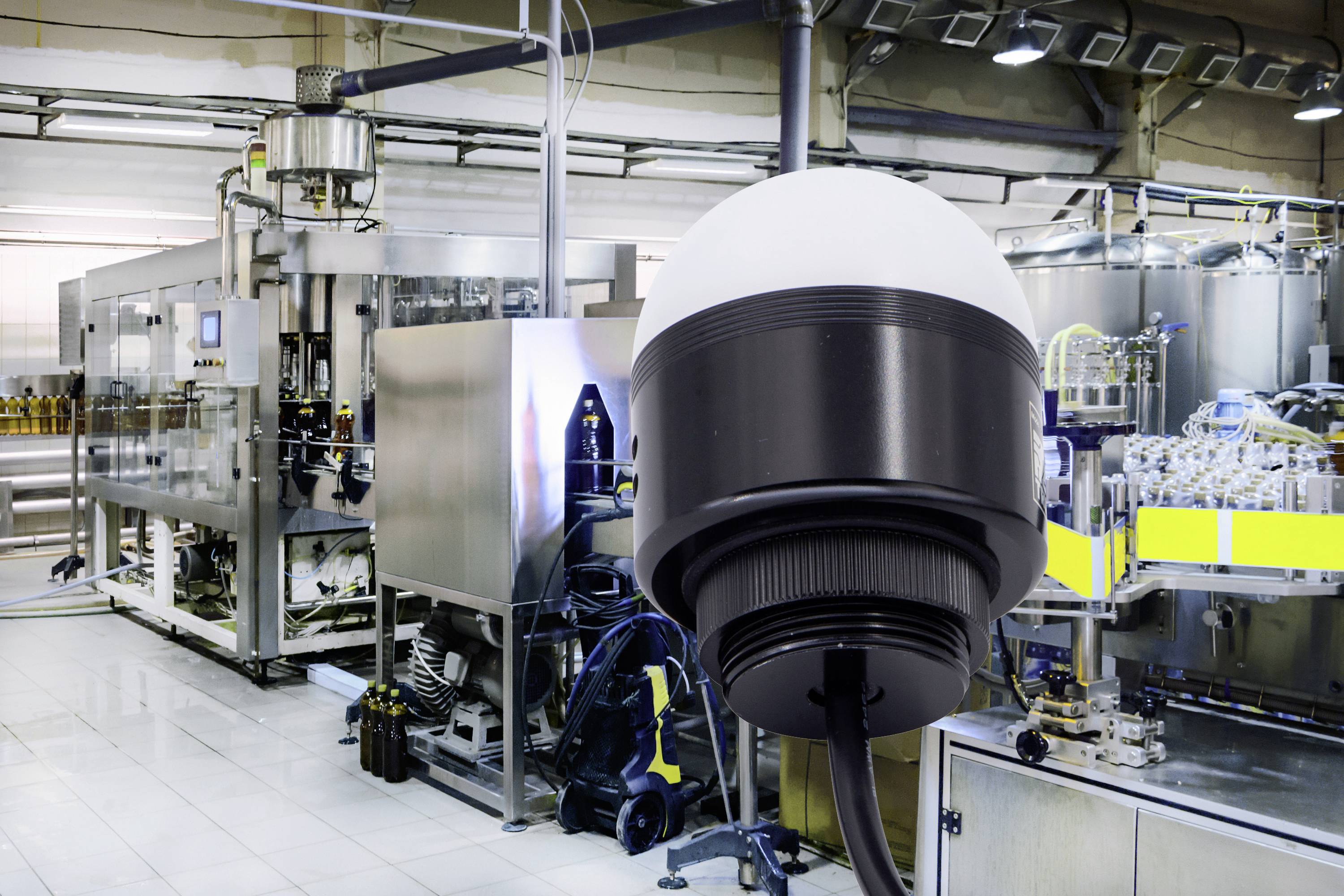 Security camera in the foreground, industrial production facility in the background with machinery and conveyor belts in a factory setting.