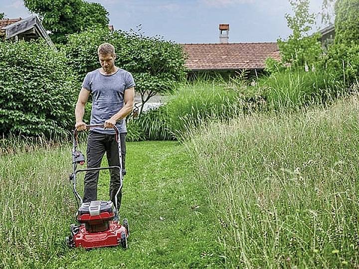 A person is mowing the lawn with a red lawnmower in a garden. Trees and a house can be seen in the background.