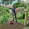 A woman is working in the garden with an electric cultivator. Flower beds and trees can be seen in the background.