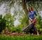 A woman in a blue top is using a leaf blower to vacuum leaves into a pile in a garden, surrounded by trees and flowers.