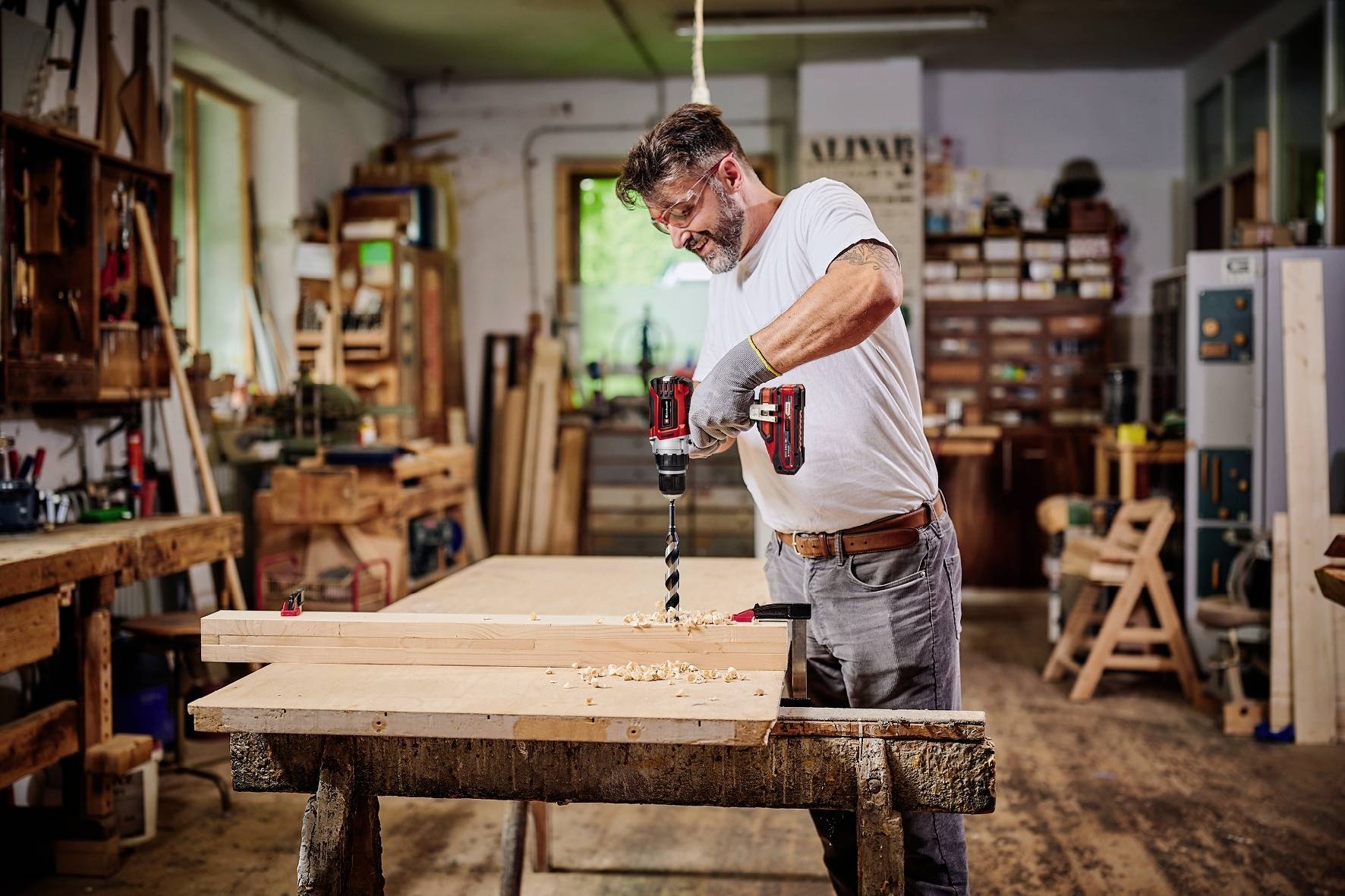 A man in a workshop is using a drill to work on a wooden board. Surrounded by tools and wood.