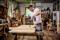A man in a workshop is using a drill to work on a wooden board. Surrounded by tools and wood.