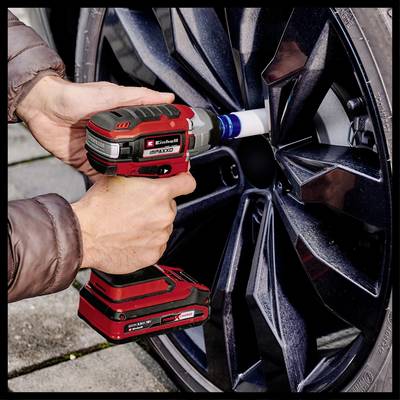 A person wearing a brown jacket is using a red cordless impact driver on the wheel nut of a black car.