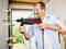 A man is drilling a hole in a wall using a drill. He is wearing safety glasses and smiling while carrying out his task.