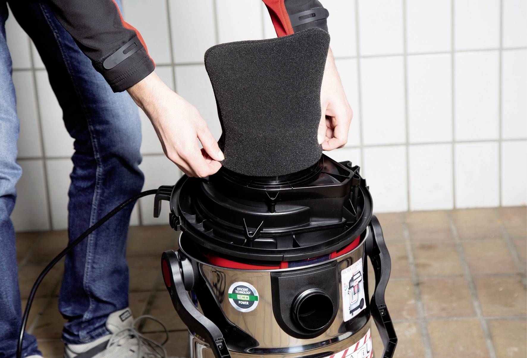 A person is replacing the air filter of a wet and dry vacuum cleaner that is standing on a tiled floor.