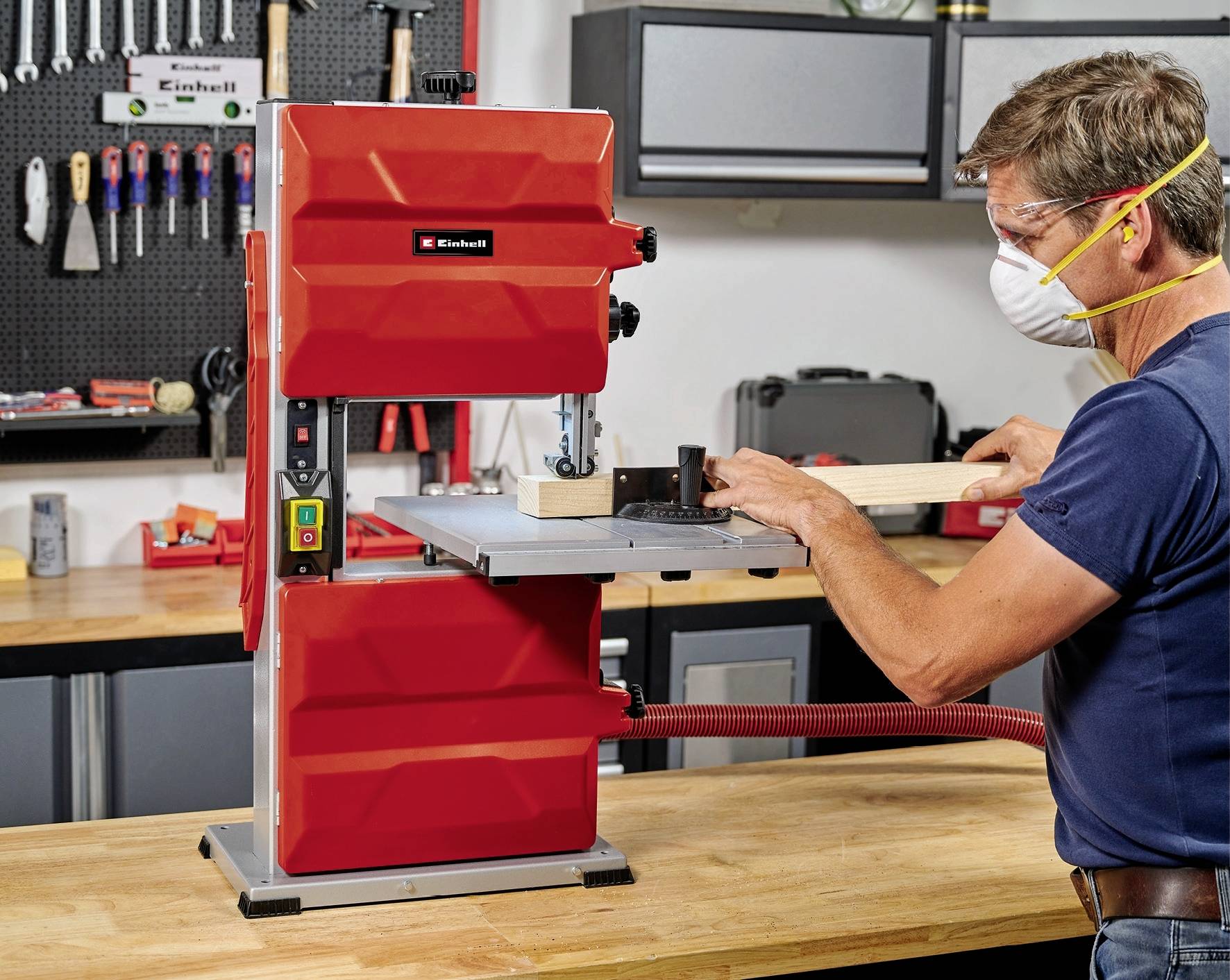 A man is wearing a protective mask and operating a red bandsaw to cut a piece of wood in a workshop.