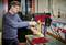 A man is working on a red pillar drill in a workshop. He is drilling a hole in a piece of wood, surrounded by tools.