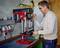 A man is operating a bench drill in a workshop. He is wearing safety glasses and holding the lever with his right hand.