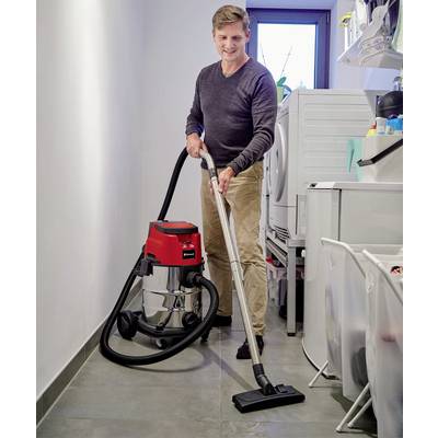 A man is hoovering the floor in a utility room. Next to him are a washing machine and various household items.