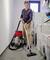 A man is hoovering the floor in a utility room. Next to him are a washing machine and various household items.