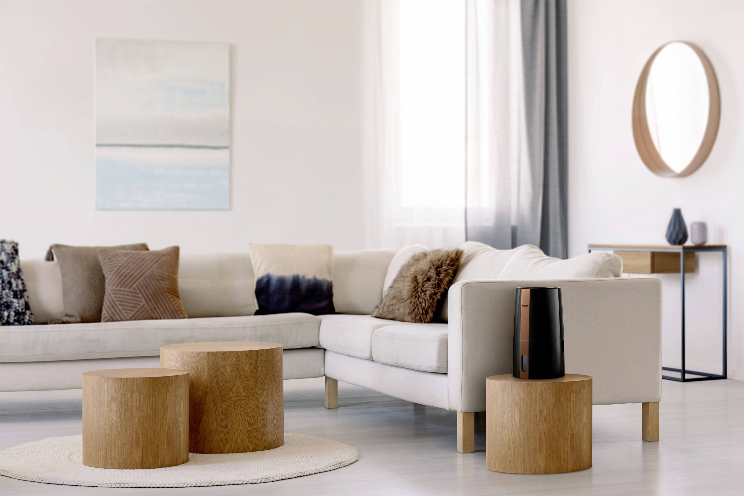 A modern living room with a light-coloured sofa, wooden tables and an air purifier placed on one of the tables. A round mirror hangs on the wall.