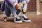 A person sitting holds a black object in front of a skateboard on a cobblestone street. Focus on shoes and hands.