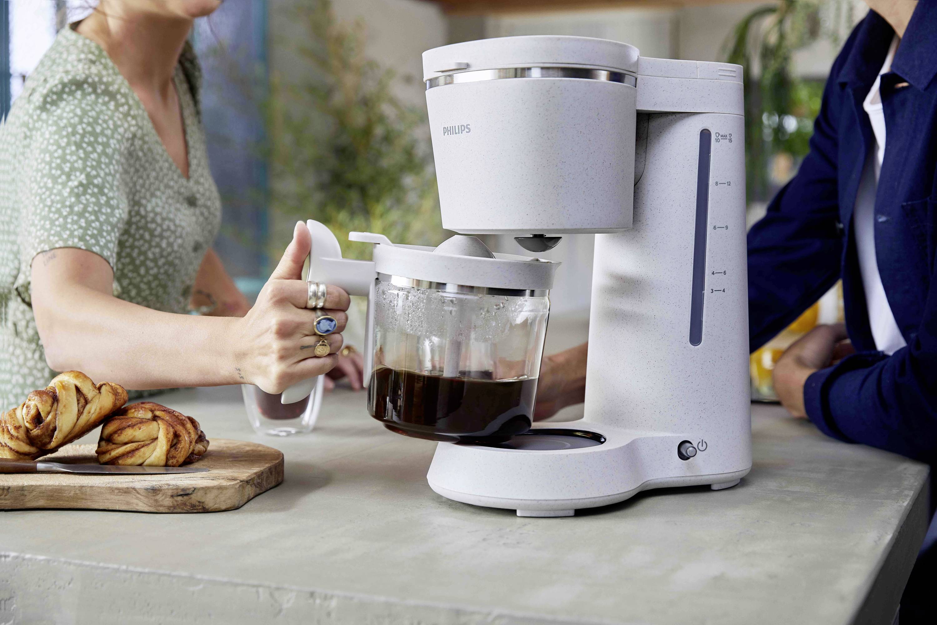 A woman is pouring coffee from a white coffee machine into a cup, with another person sitting beside her. Pastries are laid out in the foreground.