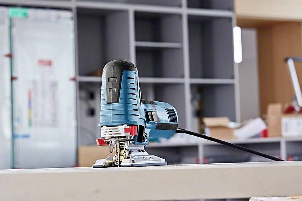 A blue jigsaw saw is lying on a workbench in a workshop. Shelves and tools are visible in the background.