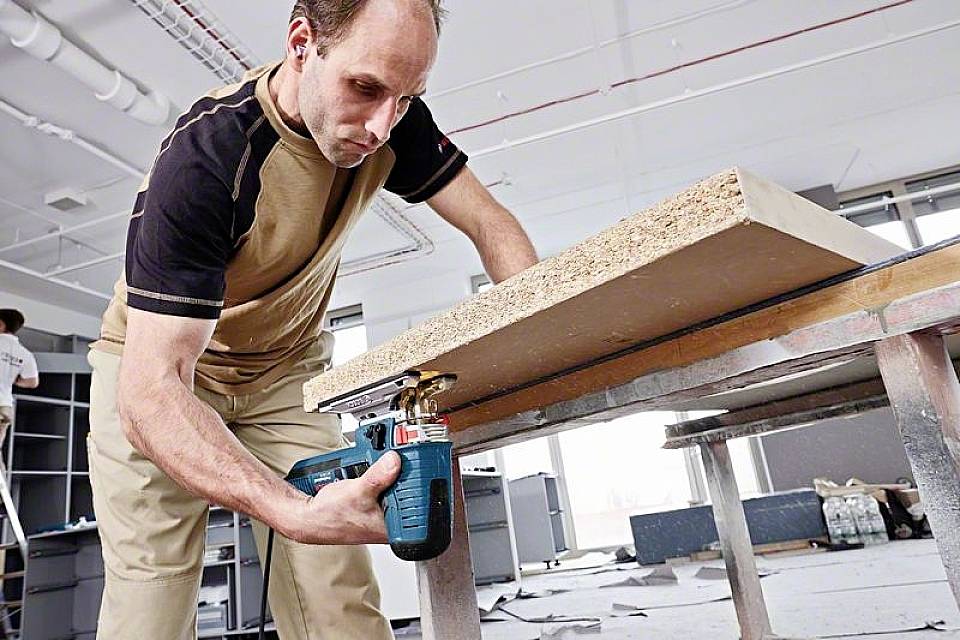 A man is using an electric jigsaw to cut a wooden board in a workshop area. Additional tools are visible in the background.