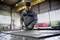 A person wearing protective equipment is grinding metal in a factory hall. Sparks are flying from the grinding wheel, and the hall is well-lit.
