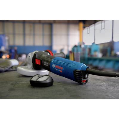 A blue angle grinder lies on a table in a workshop. A piece of metal is positioned to the left of it. Machines are blurred in the background.