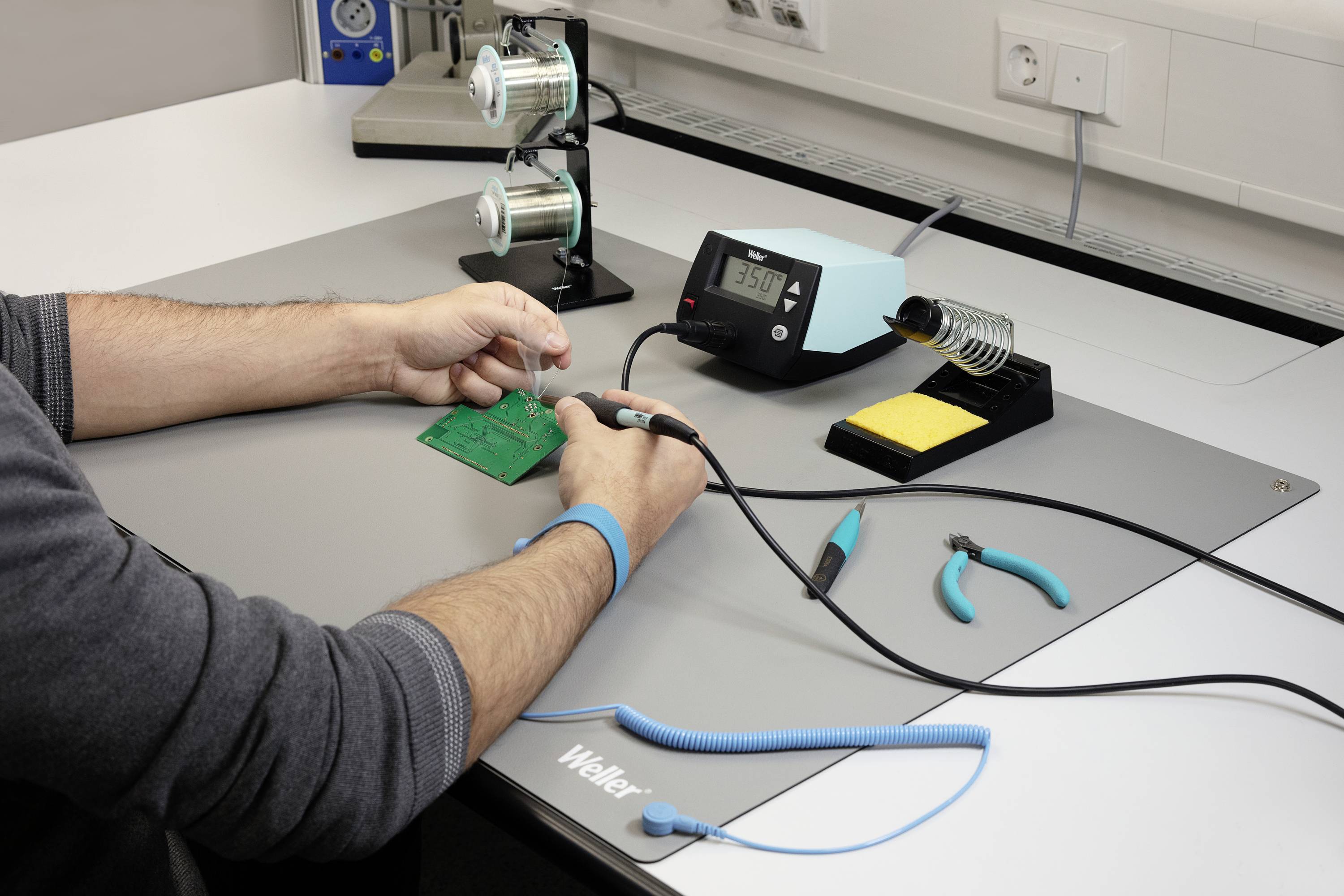 A person is soldering on a circuit board at a table with a soldering station, solder reels, and tools. Power sockets are visible in the background.