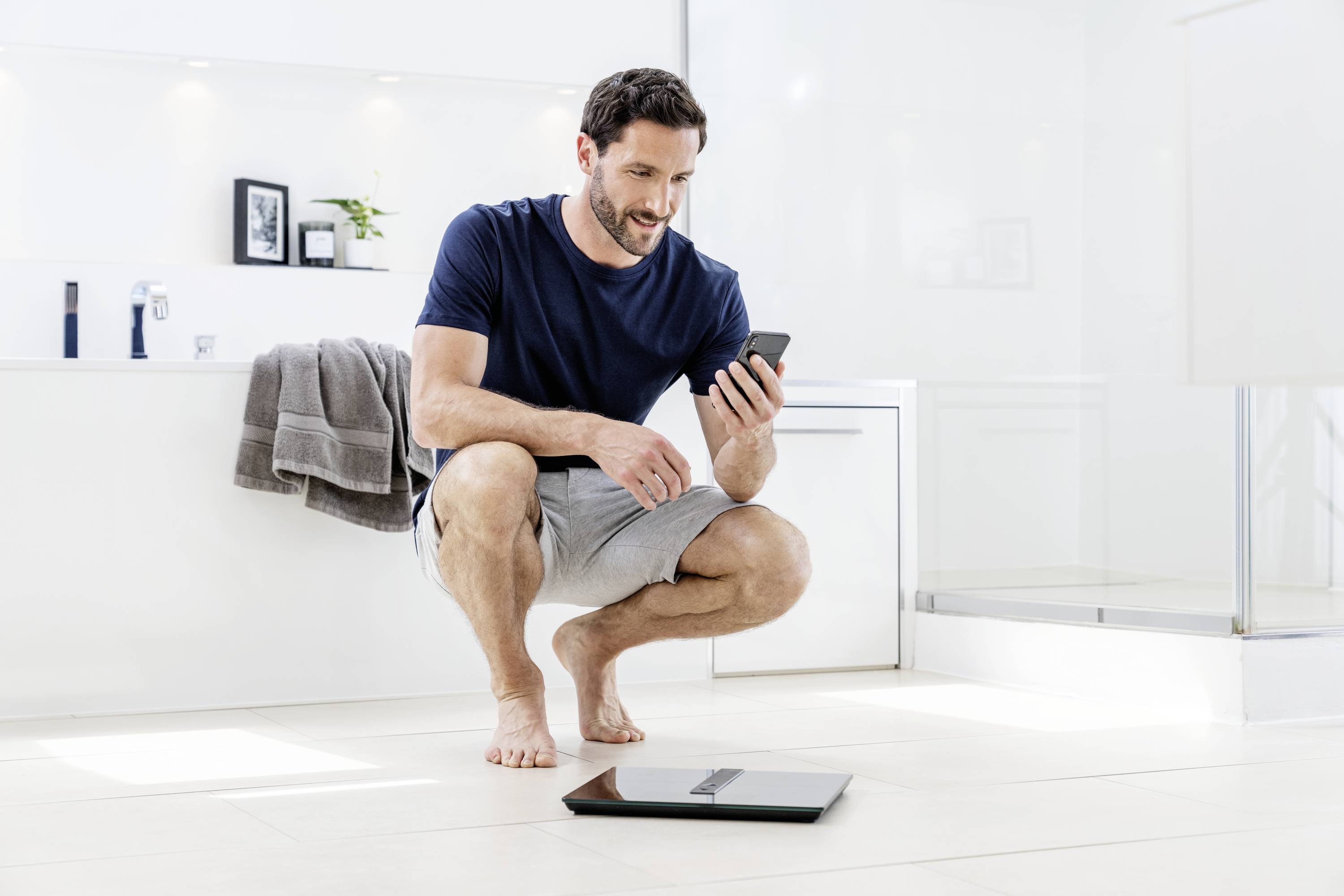 A man is sitting in a bathroom and looking at his mobile phone. A digital scales lies on the floor in front of him.