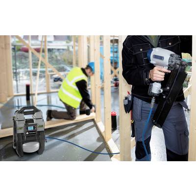 A person is working on a building site with a pneumatic nail gun. A portable compressor is in the foreground. A worker is in the background.