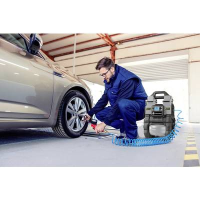 A mechanic is checking the tyre pressure of a car in a workshop. Next to him is an air compressor with an attached hose.