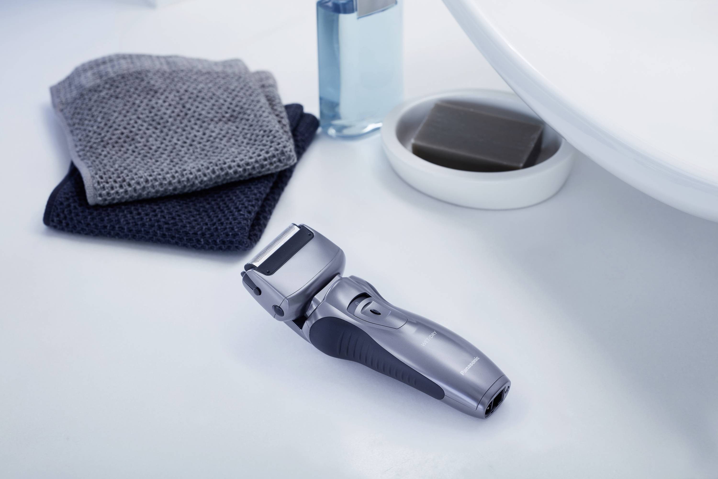 Electric shaver on a bathroom washbasin, beside grey towels, a soap dish, and a blue bottle.