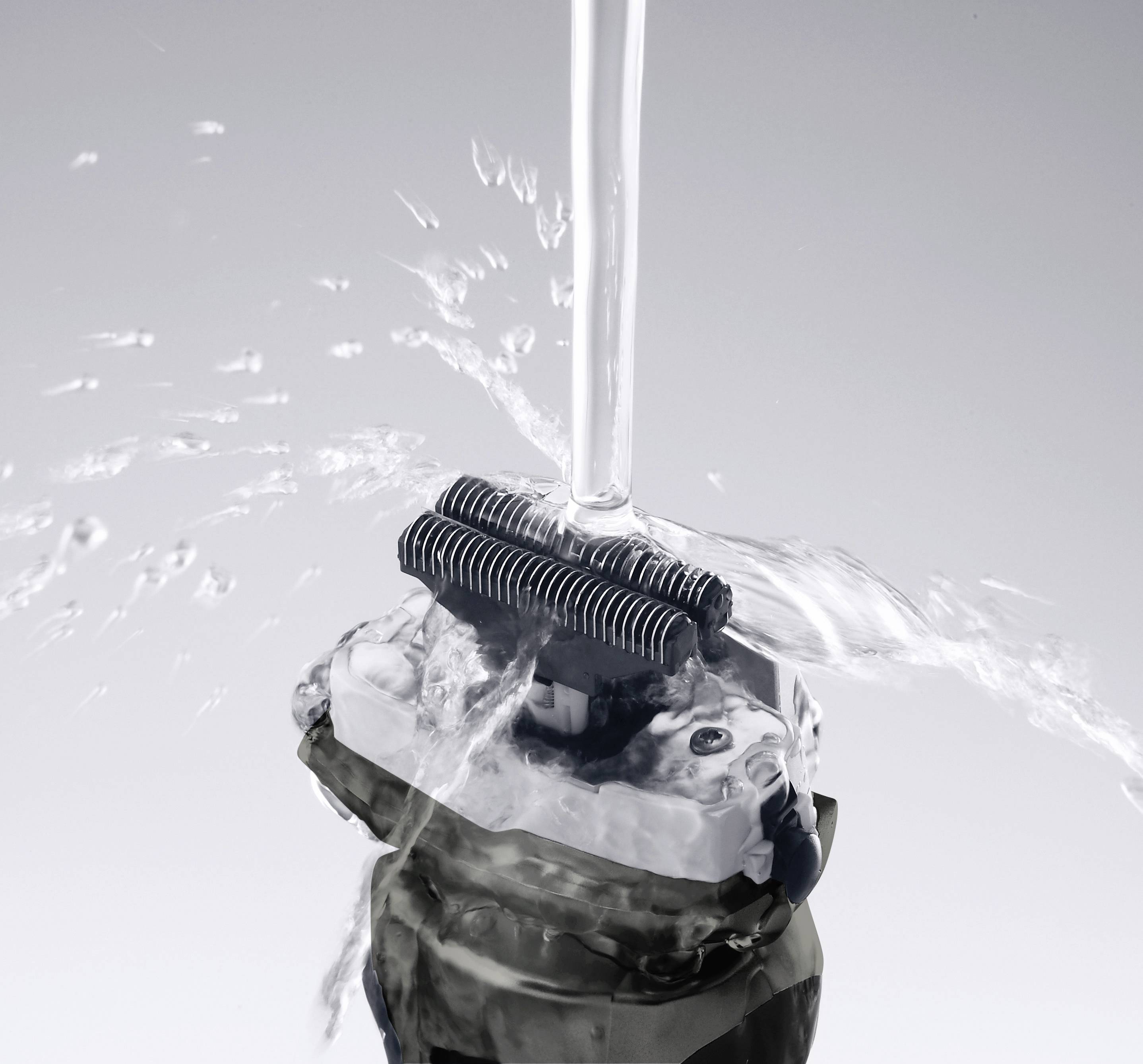 Electric razor being rinsed under a water jet. Water sprays around the blades, which are being cleaned.