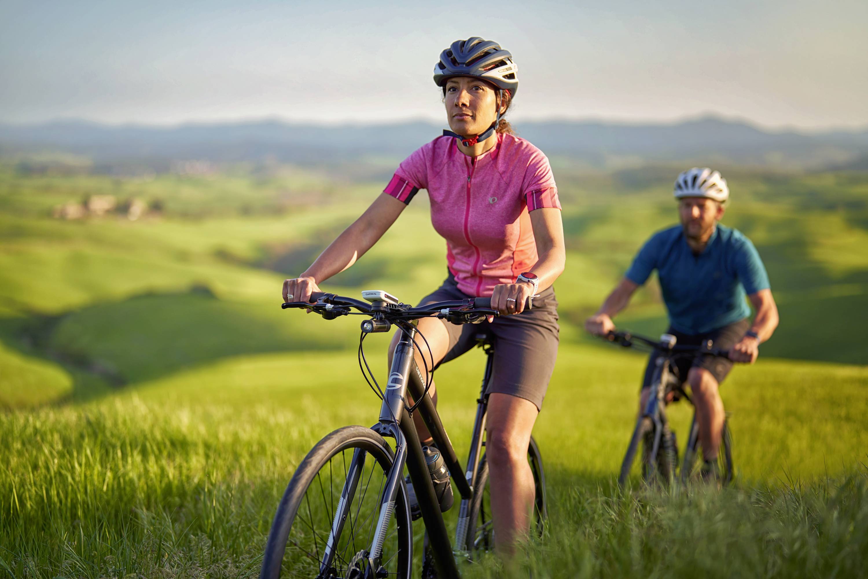 Two people are cycling through a green, hilly landscape. In the foreground, a woman wearing a red jersey.