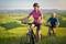 Two people are cycling through a green, hilly landscape. In the foreground, a woman wearing a red jersey.