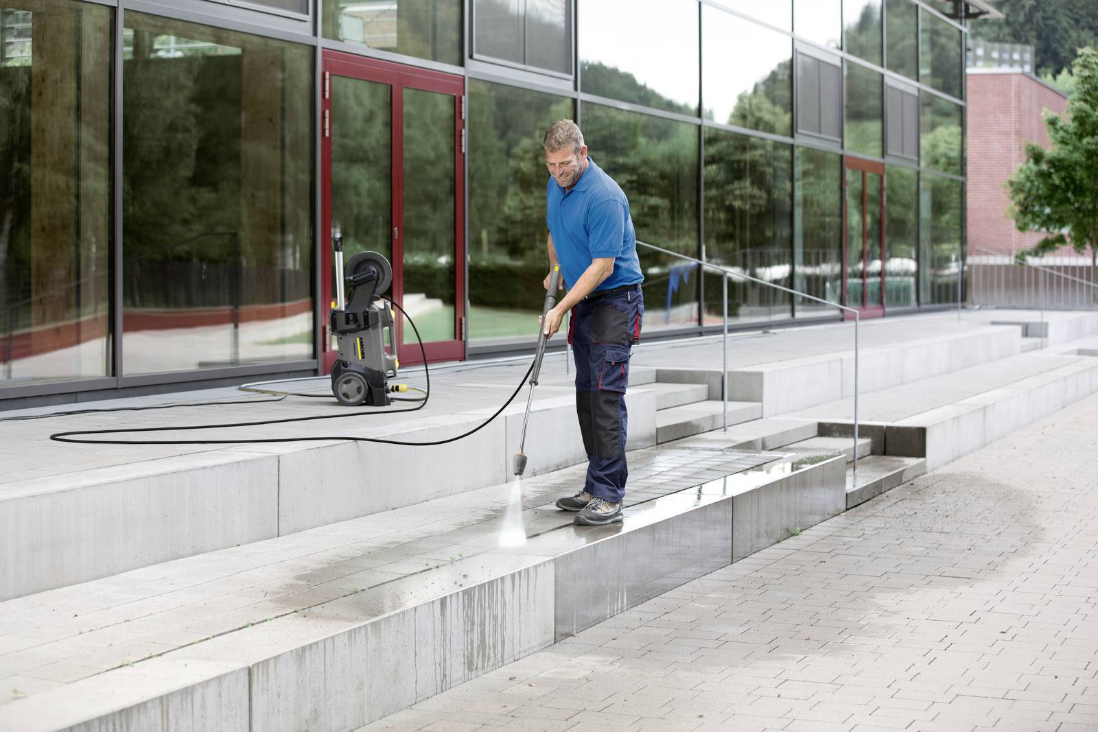A person power washes the steps of a modern building. The area appears wet and clean, with reflections visible in the glass facade.