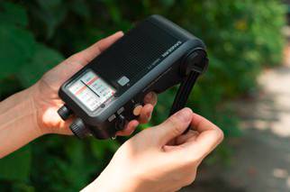 A person is holding a portable, black radio with a tuner display and rotary knobs, surrounded by green plants in the background.