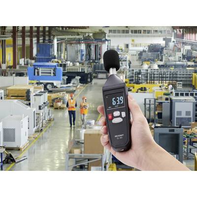 A person is holding a decibel meter showing 63.9 dB in a large factory environment with machinery and workers in the background.