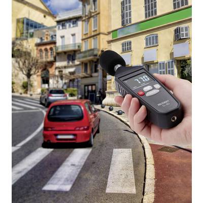 A person is measuring noise levels with a sound level metre beside a busy road with a red car passing by.
