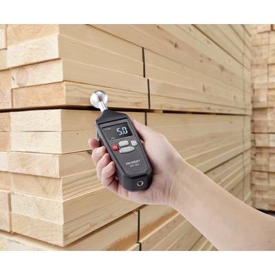 A hand holds a moisture metre in front of stacked wooden planks. The display shows a reading of 5.0.