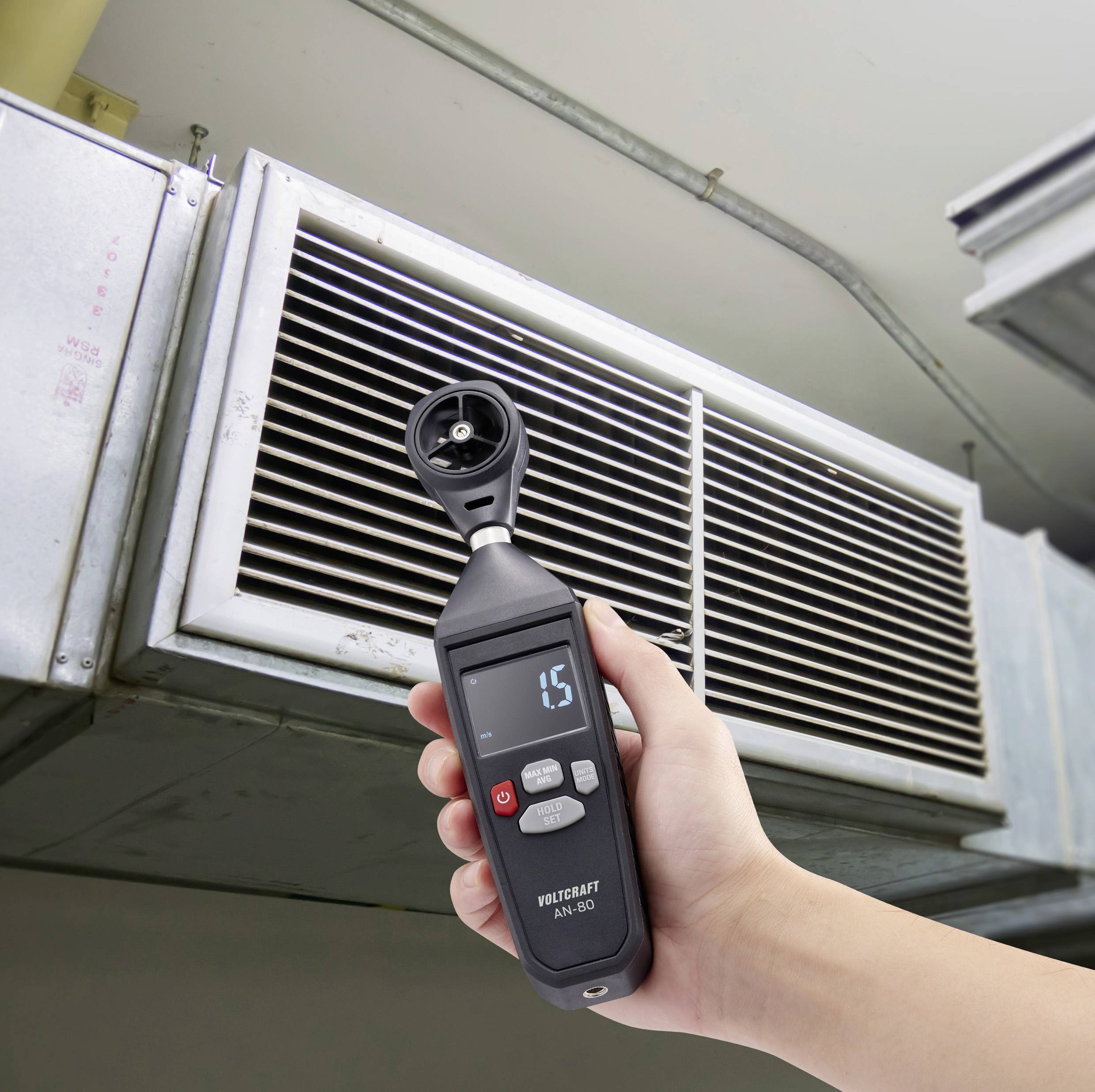 A hand holds an anemometer measuring the air velocity of a ventilation system. The display shows '1.5'.