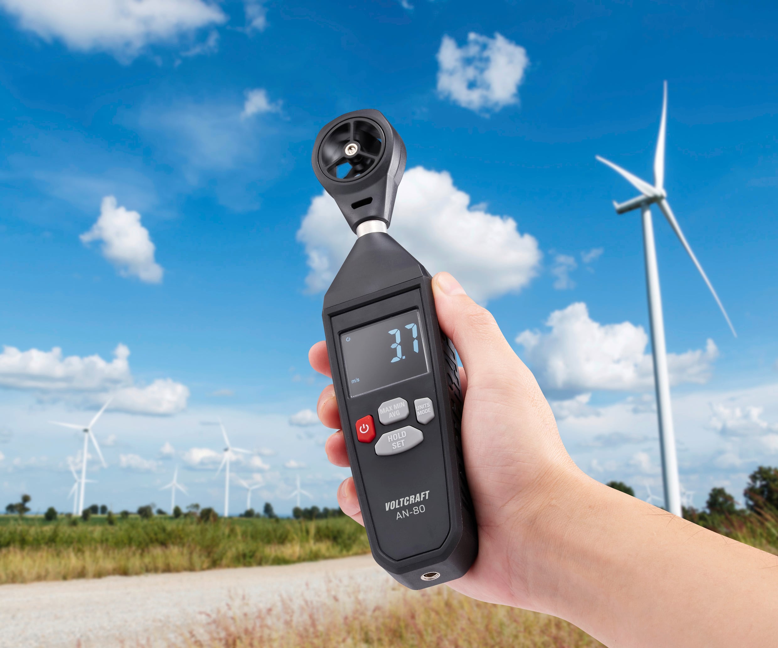 A hand is holding an anemometer displaying a wind speed of 3.7. Several wind turbines can be seen in the background.