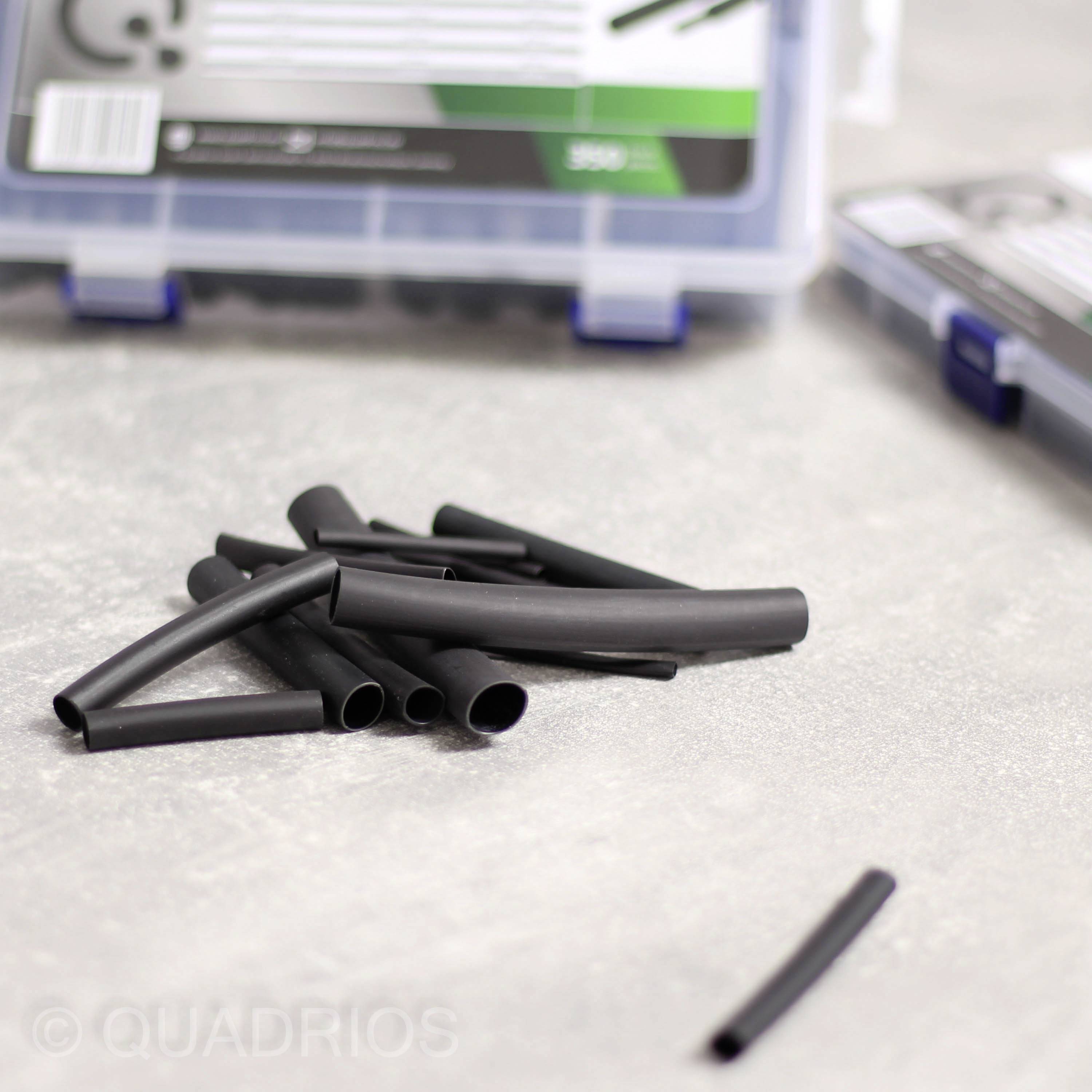 Several black heat-shrink tubes lie in front of a transparent container on a grey table. The container has a green label.