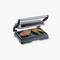 An electric contact grill with its lid open is grilling two steaks and green asparagus spears wrapped in bacon.
